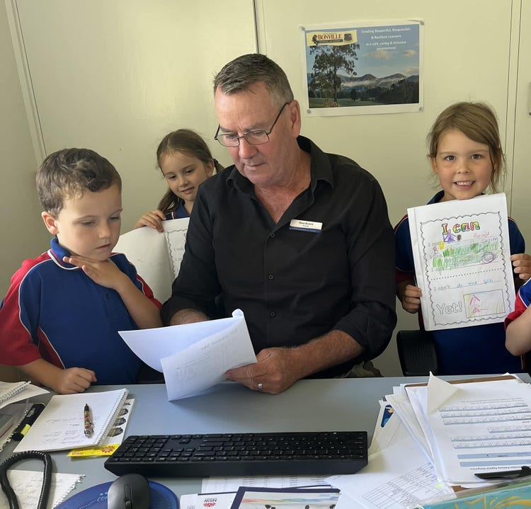The principal sitting at his desk surrounded by three kindergarten students who are showing him their writing