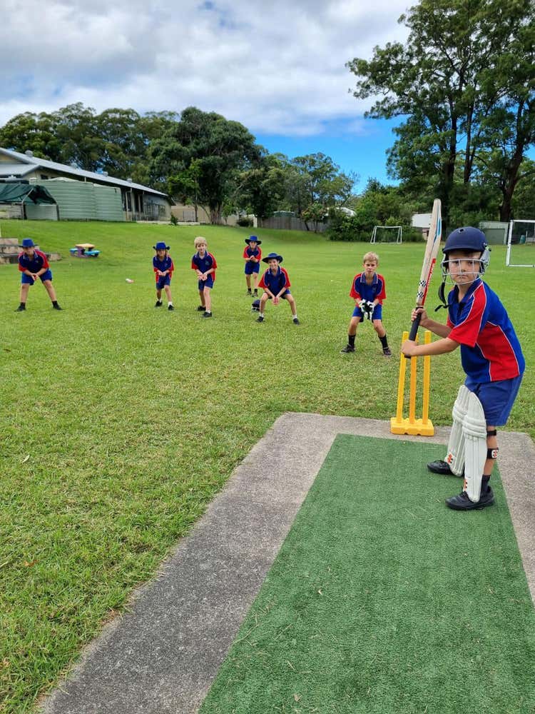 A student in cricket pads and helmet with his bat ready on the cricket pitch. Several other students are standing behind in fielding positions.