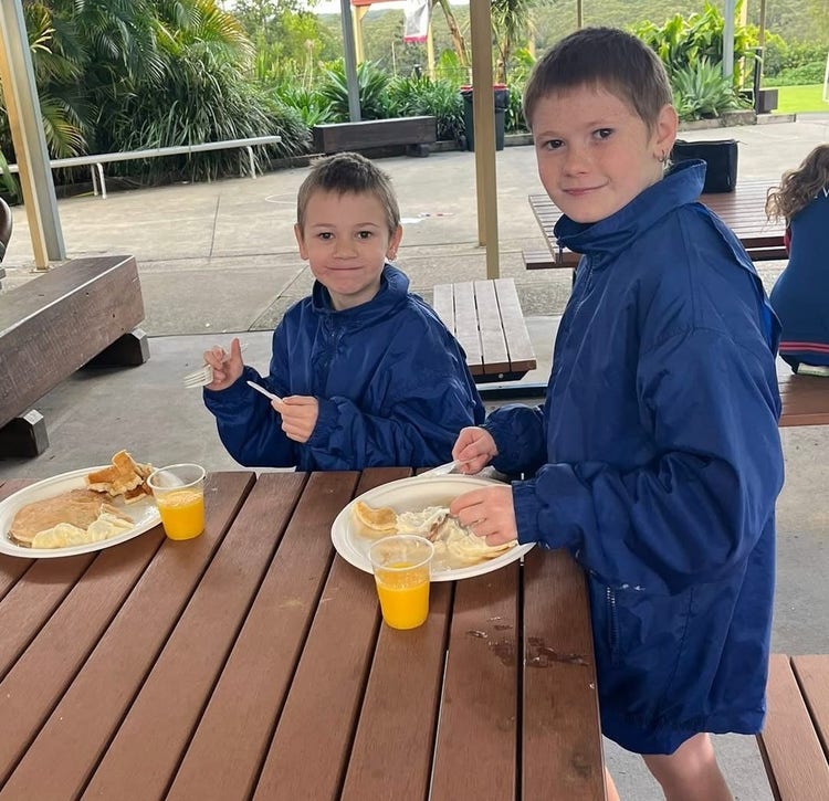 Two students at a picnic table, each with a plate of pancakes and a cup of orange juice