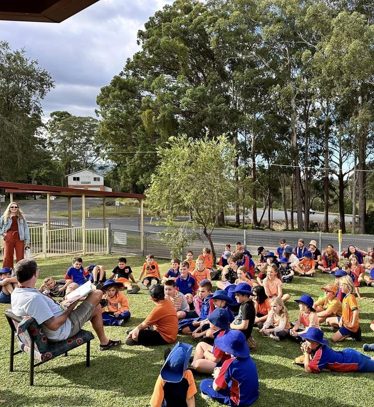 A teacher reading a story to a large group of school students on the grass