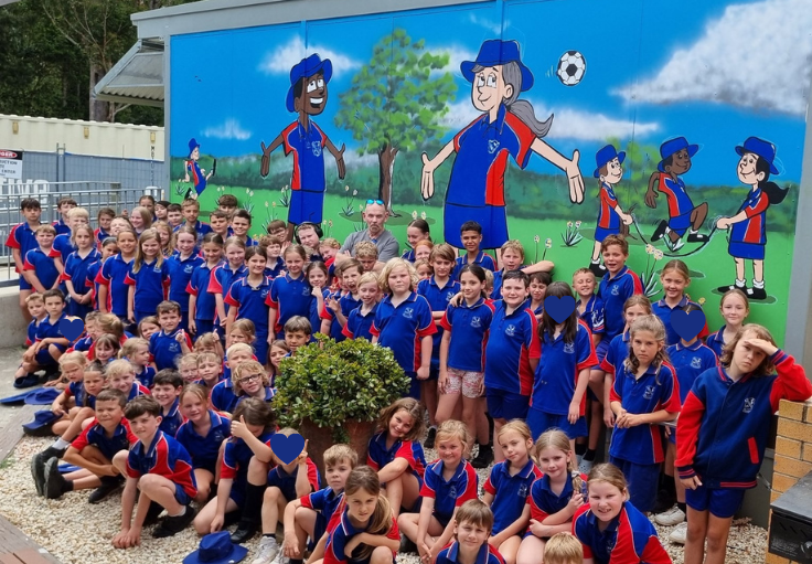 All Bonville students are sitting or standing in front of the new mural they helped to paint. The mural is a cartoon style picture of students in Bonville uniform playing on the grass.