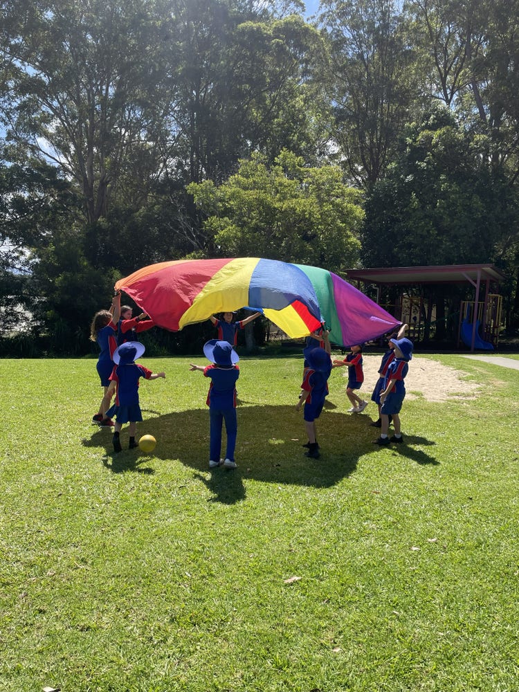 A number of kindergarten students and Year six buddies lifting up a rainbow parachute in the school playground