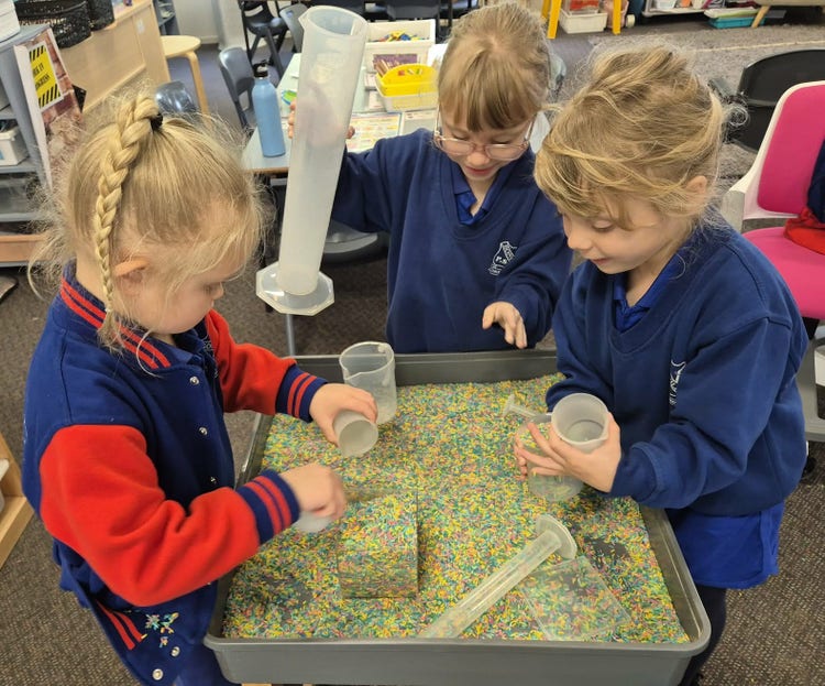 Three kindergarten students playing with coloured rice and measuring cups in a plastic tray